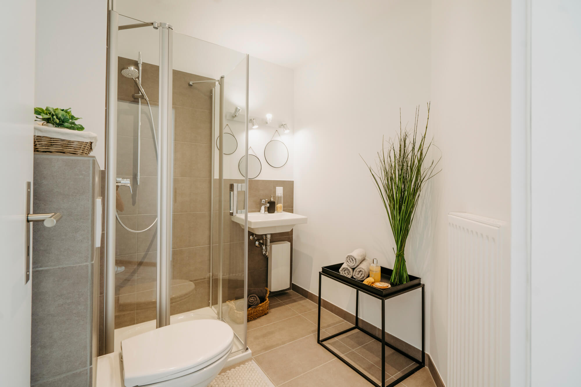 Lisa Treusch Fotografin Immobilienfotografin Nahaufnahme Badezimmer Dusche mit Glaswand drei Spiegeln an der Wand und einem schwarzen Regal im Industrial Stil mit drei gerollten Handtüchern und grüner Pflanzendekoration
